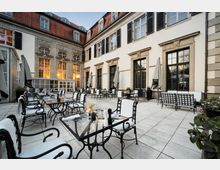 Innenhof eines historischen Gebäudes mit gepflastertem Boden, umgeben von cremefarbenen Wänden mit großen Fenstern und dekorativen Steinelementen. Metallstühle mit weißen Polstern sowie gläserne Tische und Sonnenschirme sind für Restaurantgäste aufgestellt.