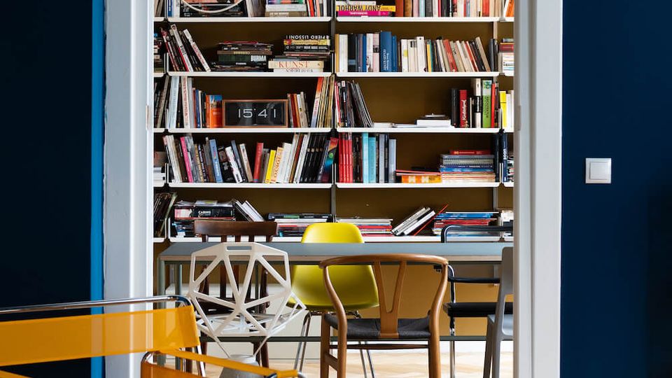 View through a white doorway into a home library or study with built-in wall-to-wall bookshelves, a central table with mixed modern chairs, and a hanging pendant light. The room has a herringbone wood floor and dark blue walls with colorful seating visible in the foreground.