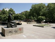 Ein öffentlicher Platz mit einer Bronzestatue auf einem Sockel im Vordergrund, daneben ein kleiner Spielplatz mit Rutschen und Sandfläche, umgeben von Bäumen und Wohngebäuden im Hintergrund. Auf dem Platz befinden sich Sitzbänke und gepflasterte Flächen.