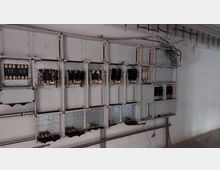 Wand mit einer Reihe leerer und teilweise beschädigter elektrischer Schaltkästen in einem einfachen, unbehandelten Raum. Einige Kabel und Leitungen hängen lose, und die Oberfläche der Wand ist schlicht und weiß.