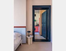 Ein Schlafzimmer mit Blick durch eine geöffnete Tür in eine Küche. Im Vordergrund ein Bett mit weißer Bettwäsche und ein kleiner Beistelltisch mit Lampe und Büchern. Die Tür hat einen dunklen Rahmen, und die Küche im Hintergrund zeigt bunte Schrankfronten in Rot, Grün und Holzoptik.