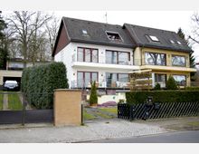 Zweistöckiges Gebäude mit Satteldach, das als Pension genutzt wird, mit Balkonen und großen Fenstern. Vor dem Haus befinden sich eine Zufahrt mit Tor, gepflegte Hecken, ein kleiner Garten und ein Schild mit der Aufschrift „Pension.“