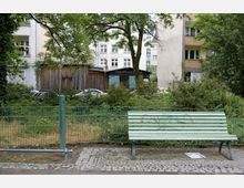 Eine Grünfläche mit einem Zaun im Vordergrund, dahinter Sträucher, ein Baum und ein hölzernes Gebäude. Im Hintergrund sind Wohnhäuser mit mehreren Stockwerken zu sehen; auf dem Pflasterweg vor dem Zaun steht eine grüne Parkbank mit Graffiti.