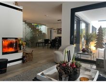 Offener, moderner Wohn- und Essbereich mit verglastem Kamin links, Holzfußboden und Treppe mit Glasgeländer im Hintergrund. Rechts führen große bodentiefe Schiebetüren auf eine Terrasse mit Pflanzen und dekorativer Beleuchtung.