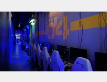 Ein schmaler Raum mit einer Reihe von Gaming-Stationen auf der rechten Seite, ausgestattet mit weißen Gaming-Stühlen, Monitoren und Kopfhörern. Die Wände sind dunkel gehalten und mit blauer sowie gelber Beleuchtung und großen Zahlen dekoriert, die futuristische Atmosphäre vermitteln.
