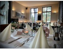 Elegantes Restaurant mit gedeckten Tischen, weißen Tischdecken, gefalteten Stoffservietten, Blumenarrangements und Kerzen. Große Fenster im Hintergrund lassen Tageslicht in den Raum.