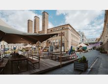 Offener Innenhof mit Holztischen und Bänken, teilweise überdacht durch ein großes Segeltuch. Im Hintergrund sind industrielle Gebäude mit hohen Schornsteinen und Containerstrukturen zu sehen, dekoriert mit bunten Bändern; die Umgebung wirkt urban mit historischem Charakter.