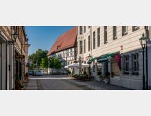 Straße mit Kopfsteinpflaster, flankiert von Fachwerkhäusern auf der linken Seite und einem historischen Gebäude mit heller Fassade auf der rechten Seite; Außengastronomie mit Sonnenschirmen und Sitzplätzen, umgeben von Altstadtbeleuchtung. Im Hintergrund Bäume und parkende Autos.