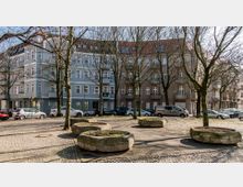 Straßenszene mit mehrgeschossigen Wohnhäusern im Hintergrund, teils blau und teils beige gestrichen, mit Balkonen und vielen Fenstern. Im Vordergrund stehen kahle Bäume sowie große, runde Pflanzkübel auf einem gepflasterten Platz, umgeben von parkenden Autos.
