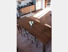 Esszimmer mit einem langen Holztisch und mehreren Stühlen aus Metall. Im Hintergrund eine offene Küche mit Holzschränken, einer schwarzen Arbeitsfläche, einem Edelstahl-Kühlschrank und gemauerten Details.