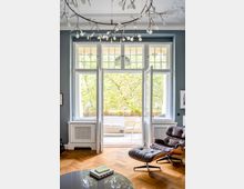 Wohnzimmer mit großen weißen Doppelfenstern und Blick auf einen Balkon mit Sitzmöbeln und Pflanzen. Ein moderner Ledersessel mit Fußhocker steht auf einem Holzboden, darüber hängt eine filigrane Deckenleuchte mit Zweigmuster.