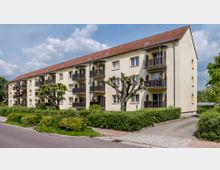 Mehrfamilienhaus mit drei Stockwerken und gelber Fassade, mehreren Balkonen und einem roten Satteldach. Davor befinden sich gepflegte Büsche, ein baumgesäumter Gehweg und eine gepflasterte Zufahrt.