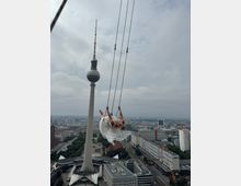 Eine Frau in einem weißen Kleid schaukelt hoch über der Stadt von einer Schaukel, mit dem Berliner Fernsehturm im Hintergrund. Im unteren Bereich sind Gebäude und Sehenswürdigkeiten von Berlin, wie die Marienkirche und Teile des Alexanderplatzes, zu sehen.