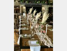 Langgedeckter Holztisch in einem rustikalen Raum mit Steinwand und Kamin, dekoriert mit beigefarbenen Stoffläufern, Trockenblumen, rosa Rosen, Kerzenhaltern und eingedecktem Geschirr. Im Hintergrund sind Holzstühle und ein Kamin mit Dekoration sichtbar.