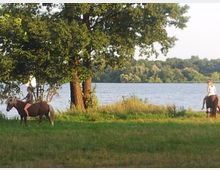 Zwei Personen reiten auf Ponys auf einer grünen Wiese, die von Bäumen umgeben ist, mit einem See und bewaldetem Ufer im Hintergrund. Die Szene findet in einer natürlichen Umgebung bei Tageslicht statt.