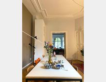 Wohnzimmer oder Essbereich mit einem langen weißen Tisch und Holzstühlen, darauf eine Vase mit bunten Blumen, Schalen und Kerzen. Im Hintergrund ist eine geöffnete Doppeltür, die in einen weiteren Raum mit einem großen Fenster und Sofa führt. Die hohen Decken zieren dekorative Stuckelemente, und der Boden ist mit hellem Parkett und einem Teppich gestaltet.