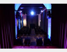 Blick von der Bühne in einen kleinen Veranstaltungssaal mit Reihen aus dunklen Stühlen und einem Mittelgang. Dunkle Vorhänge, seitliche Säulen und blau-violette Bühnenbeleuchtung sowie Lautsprecheranlagen prägen den Raum.