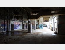 Eine verlassene, heruntergekommene Fabrikhalle mit beschädigten Wänden, herunterhängenden Deckenstrukturen und Graffiti bedeckten Flächen. Der Boden ist mit Schutt bedeckt, während das Tageslicht durch große Fensterflächen einfällt.