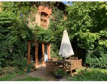 Backsteinfassade eines Hauses mit großen bodentiefen Holzfenstern, umgeben von dichtem Efeubewuchs. Im Vordergrund steht eine Terrasse mit hölzernem Gartentisch, Stühlen und einem Sonnenschirm, umgeben von Grünpflanzen und Blumen.