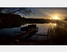 Sonnenuntergang an einem stillen See mit einem hölzernen Steg, der zu einem kleinen Boot führt. Im Vordergrund sind Schilf und Äste zu sehen, während der Himmel und die Wasseroberfläche in warmem Licht leuchten.