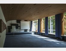 Raum mit modernem Design für Präsentationen oder Veranstaltungen, ausgestattet mit einer hölzernen Lamellendecke, großem Projektionsbildschirm, Flipchart und blauen Vorhängen vor großen Fenstern, die Tageslicht hereinlassen. Im Hintergrund ist ein erhöhtes Podium vor einer dekorativen Wandinstallation zu sehen.