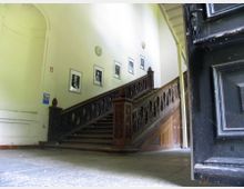 Holztreppe mit kunstvoll geschnitztem Geländer in einem historischen Gebäude. An den Wänden hängen Schwarz-Weiß-Porträts, und im Vordergrund ist eine geöffnete dunkle Tür zu sehen.