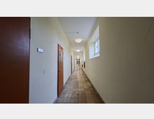 Langer, schmaler Innenflur mit hellem Anstrich, gefliestem Boden und einer Reihe von Türen auf der linken Seite. Rechts befinden sich kleine Fenster und Wandnischen, die Decke ist mit runden Leuchten entlang des Gangs ausgestattet.