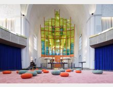 Innenraum einer modernen Kirche mit hohen weißen Wänden und farbigem Glasdesign im Altarbereich in Grün- und Blautönen, gerahmt von orangen Strukturen. Auf dem rosafarbenen Teppichboden liegen mehrere runde Sitzkissen in Orange und Grau, umgeben von blauen Vorhängen und schlichten architektonischen Elementen.