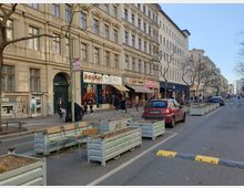 Straßenansicht einer belebten Einkaufsstraße mit Altbaufassaden, Geschäften im Erdgeschoss, geparkten Autos und Sitzbänken mit Pflanzkästen entlang der Straße. Einige Passanten sind auf dem Gehweg unterwegs, während kahle Bäume die Szenerie säumen.
