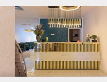 Modernes Empfangsbereich einer Einrichtung mit einem goldfarbenen Tresen mit Marmorakzenten, einer Vase mit Blumen darauf, und einer dekorativen Hängelampe. Im Hintergrund sind eine grüne Wand, eine offene Tür und mehrere Sitzecken mit dunkelgrünen Polstermöbeln zu sehen.