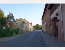 Straße in einer Kleinstadt mit einfachen Wohngebäuden auf der linken Seite und einem Ziegelbau mit torartigem Eingang auf der rechten Seite. Im Hintergrund sind Bäume und parkende Fahrzeuge sichtbar.