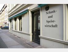 Fassade eines Restaurants mit der Aufschrift „Schank- und Speisewirtschaft“ in schwarzer Schrift. Das Gebäude hat helle Wände, große Fenster mit grünen Markisen und eine dunkle Tür; links daneben verläuft eine gepflasterte Gehsteigfläche in einer städtischen Umgebung mit weiteren Gebäuden im Hintergrund.