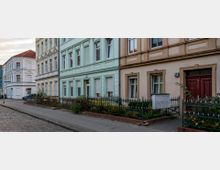 Straßenszene mit Reihen von historischen Wohnhäusern in Pastellfarben, darunter mintgrüne und beigefarbene Fassaden mit klassischen Zierleisten. Im Vordergrund befinden sich kleine Vorgärten mit Blumen und Metallzäunen, daneben ein mit Kopfsteinpflaster und Gehweg ausgestatteter Straßenabschnitt.