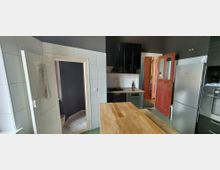 Moderne Küche mit weißen Wandfliesen und dunkelgrauen Wänden, schwarzer Hängeschrankzeile, Spüle und integrierter Spülmaschine sowie großem Holztisch im Vordergrund. Rechts stehen ein hoher Kühlschrank und ein Backofen; offene Türen führen in einen Flur und einen angrenzenden Raum.