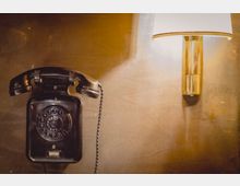 Schwarzes Wählscheibentelefon mit einem Hörer, das an einer braunen Wand hängt, daneben eine Wandlampe mit goldenem Sockel und weißem Lampenschirm.