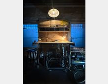 Werkstatt- oder Garageninnenraum mit hölzerner Werkbank und Regal, beleuchtet von einer hängenden Lampe und einer kleinen Arbeitsleuchte. Links und rechts befinden sich zwei Fenster; unter der Werkbank stehen Maschinen und Kabel, daneben ein großer Kompressor.