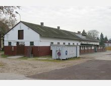 Ein langes landwirtschaftliches Gebäude mit weiß gestrichenen Wänden und einer rotbraunen unteren Hälfte, Flachdachfenstern und schrägem Dach. Im Vordergrund stehen ein großer Metallcontainer und ein "Parken verboten"-Schild, umgeben von einer unbefestigten Fläche und seitlichen Bäumen.
