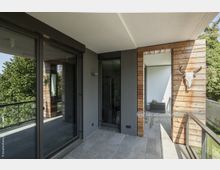 Moderne Terrasse mit grauem Boden aus Steinfliesen, einer verglasten Schiebetür sowie Holzwandverkleidungen, an der ein Tier-Schädel mit Hörnern hängt. Im Hintergrund sind grüne Bäume und ein Glasgeländer sichtbar.