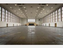 Ein großer, leerer Innenraum mit hohen Decken, rechteckigen Deckenleuchten und einer Wand aus hohen Fenstern mit braunen Rahmen. Der Raum hat eine dunkle Bodenfläche mit sichtbaren Platten, seitliche Wände mit Türen und eine Treppenanlage in der Mitte der Rückwand.