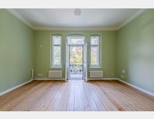 Leerer Wohnraum mit hellgrünen Wänden, hohen Holz-Dielenböden und drei hohen Fenstern, in der Mitte eine geöffnete Balkontür mit schmiedeeisernem Geländer. Unter den seitlichen Fenstern stehen Heizkörper; die Decke ist weiß mit einfachem Deckenanschluss und zentraler Leuchte.