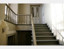 Treppe in einem alten Gebäude mit einem Geländer aus hell lackierten Holzbalustern, abgenutzten Wänden und einer weißen Doppeltür oben am Treppenabsatz. Ein Fenster wirft Licht- und Schattenmuster auf die Wand.