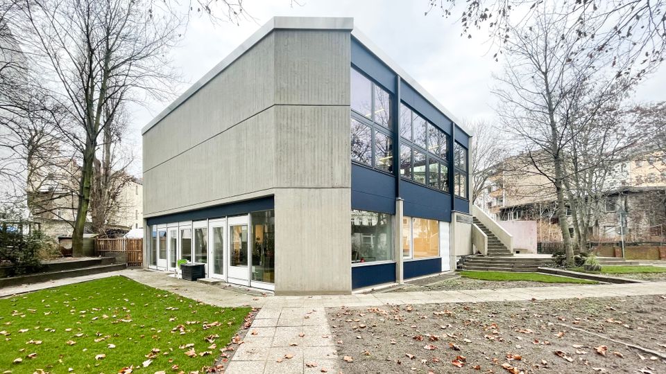 Zweistöckiges, modernes Gebäude aus Sichtbeton mit dunkelblauen Fassadenbändern und großen Fensterflächen, umgeben von kahlen Bäumen. Davor liegen gepflasterte Wege, Rasenflächen und ein Außenbereich mit Treppe und kleinen Terrassenstufen.