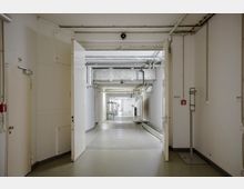 Ein langer, weiß gestrichener Gang in einem Gebäude mit sichtbaren Lüftungsrohren an der Decke, offenen Türen und einem glatten grauen Boden. Im Hintergrund stehen Regale und Tische entlang der Wände.