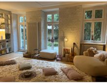 Gemütliches Wohnzimmer mit großen Fenstern und weiß gestrichenen Ziegelwänden. Der Raum ist mit einem flauschigen Teppich, Kissen, einer Gitarre, weichen Sitzmöbeln und Regalen dekoriert; durch die Fenster ist ein Garten mit Bambuspflanzen sichtbar.