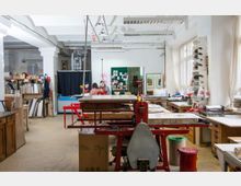 Heller Arbeitsraum mit einer Druckwerkstatt, ausgestattet mit Druckmaschinen, Arbeitstischen, Regalen voller Materialien und großen Fenstern, die natürliches Licht hereinlassen. Im Hintergrund sind zwei Personen bei der Arbeit an einem Tisch zu sehen.