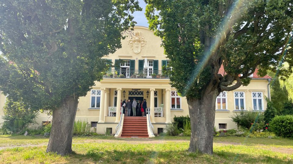 Helles, herrschaftliches Landhaus mit Säulenportal, breiter Freitreppe und Balkon mit grünen Fensterläden, vor der Fassade ein Wappenrelief. Das Gebäude liegt in einem parkartigen Garten mit großen Bäumen, Rasenfläche und Sträuchern.