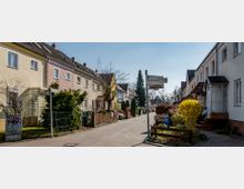 Eine ruhige Wohnstraße mit zweistöckigen Reihenhäusern in hellen Pastellfarben, vorgelagerten Gärten und einem Gehweg. Ein Wegweiser am Straßenrand zeigt Ortsnamen an, und im Hintergrund sind Bäume und ein weiteres Gebäude sichtbar.