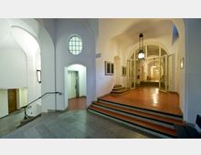 Ein historischer Innenraum mit gewölbten Decken und weißen Wänden, ausgestattet mit mehreren Treppenstufen, roten Böden und einem Aufzugsschacht auf der linken Seite. Im Hintergrund führen offene Glastüren in einen weiteren Bereich, beleuchtet von hängenden Laternen.