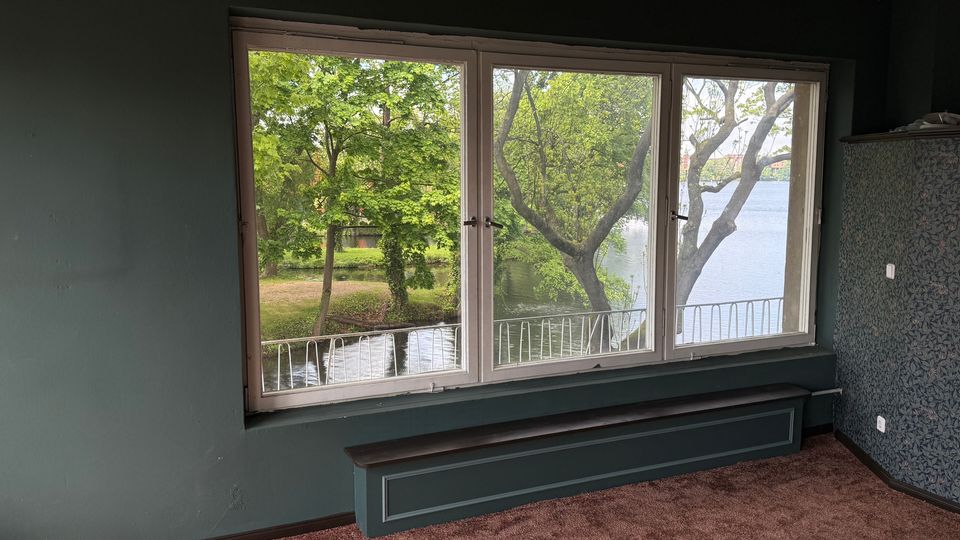 Innenraum eines leerstehenden Zimmers mit dunkelgrünen Wänden, braunem Teppichboden und großem dreiteiligem Fenster mit niedrigem Heizkörper darunter. Durch das Fenster sieht man einen Balkon mit Geländer sowie Bäume und einen See oder Fluss direkt davor.
