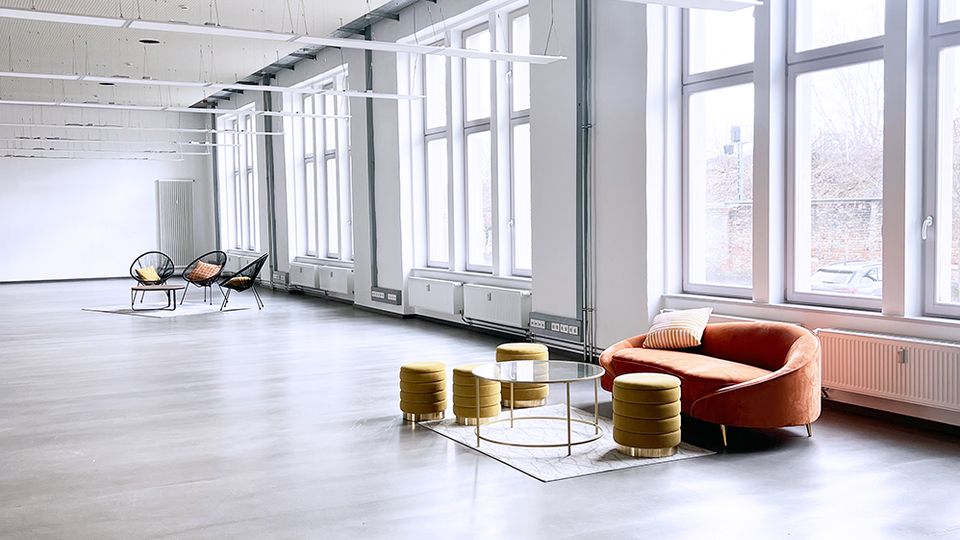 Ein modernes, helles Loft mit hohen Decken, großflächigen Fenstern und grauem Betonboden. Im Vordergrund eine Sitzecke mit einem orangefarbenen Sessel, gelben Hockern und einem Glastisch, im Hintergrund minimalistische Möbel.